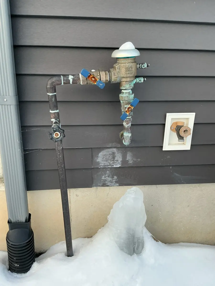 Outdoor irrigation backflow device on a house wall with a column of ice built up below it after a freeze