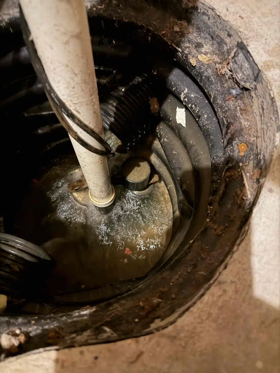 Open sump pit with an older submersible sump pump, tether float and discharge pipe inside the basin