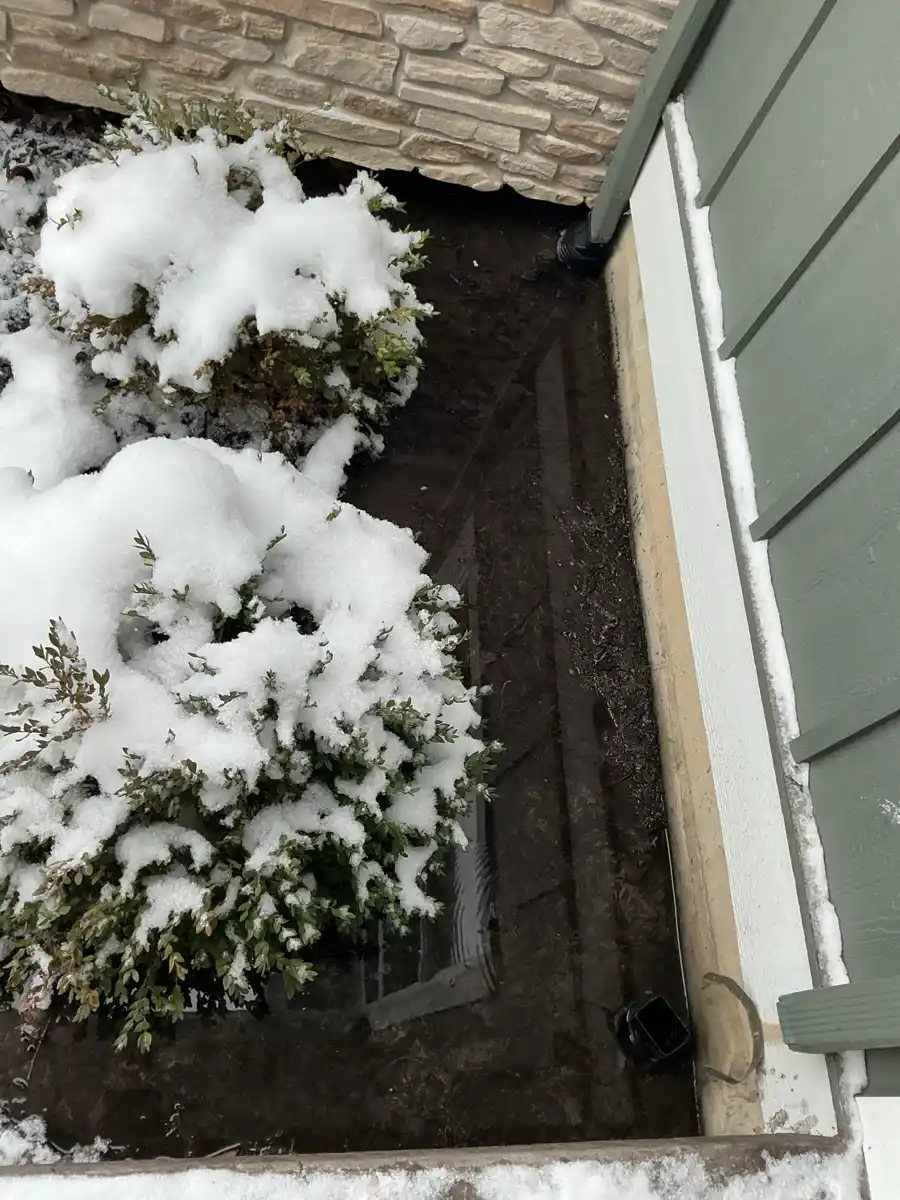 Sump pump discharge point in a snow covered bed alongside a house in winter