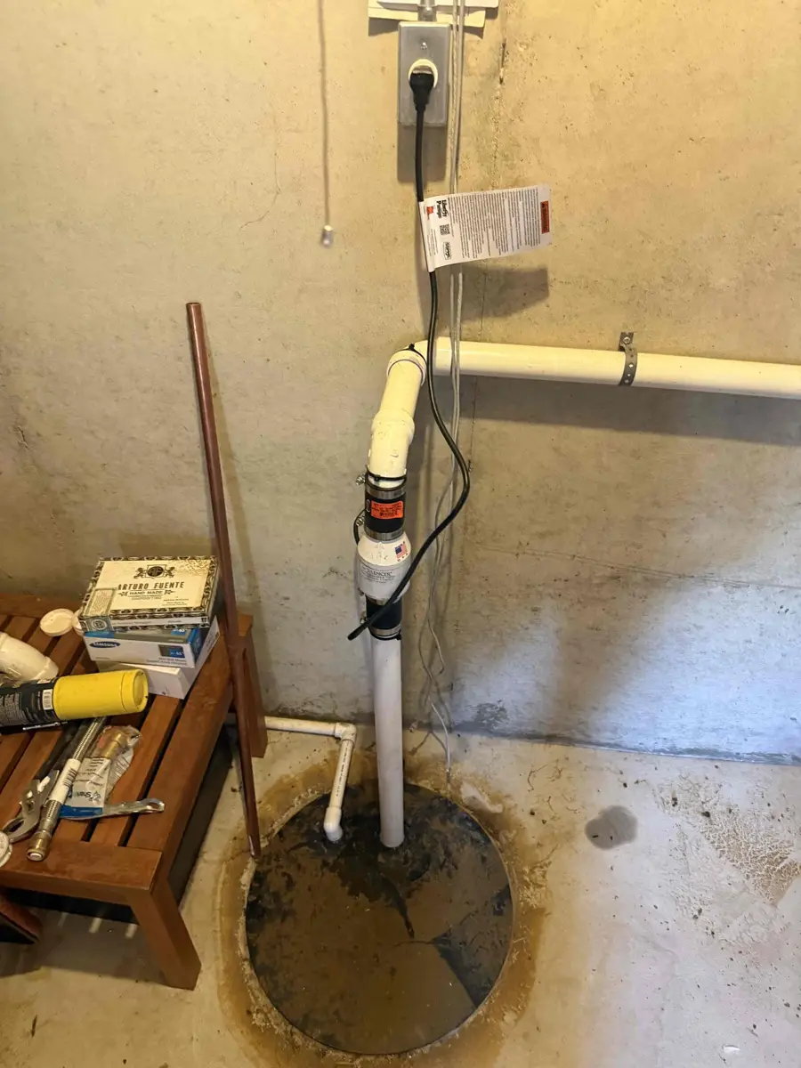 Sump pump power cord plugged into a dedicated basement outlet above the pit with discharge piping