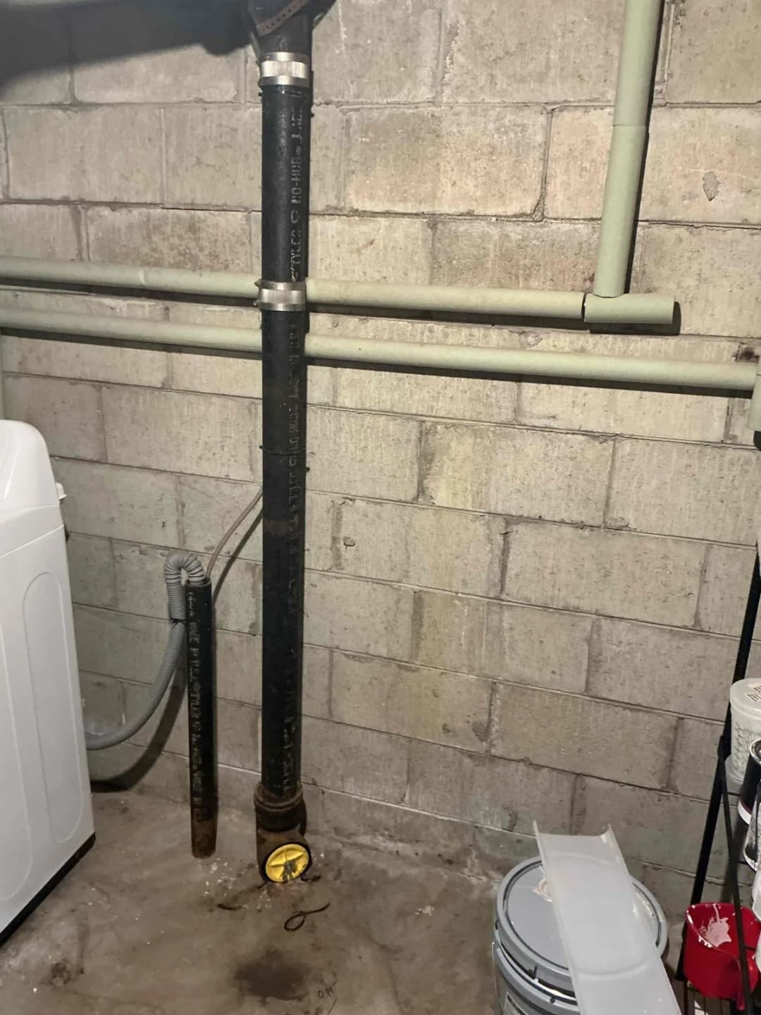 Original black cast iron main stack in a basement, with a cleanout plug near the floor and branch drains overhead