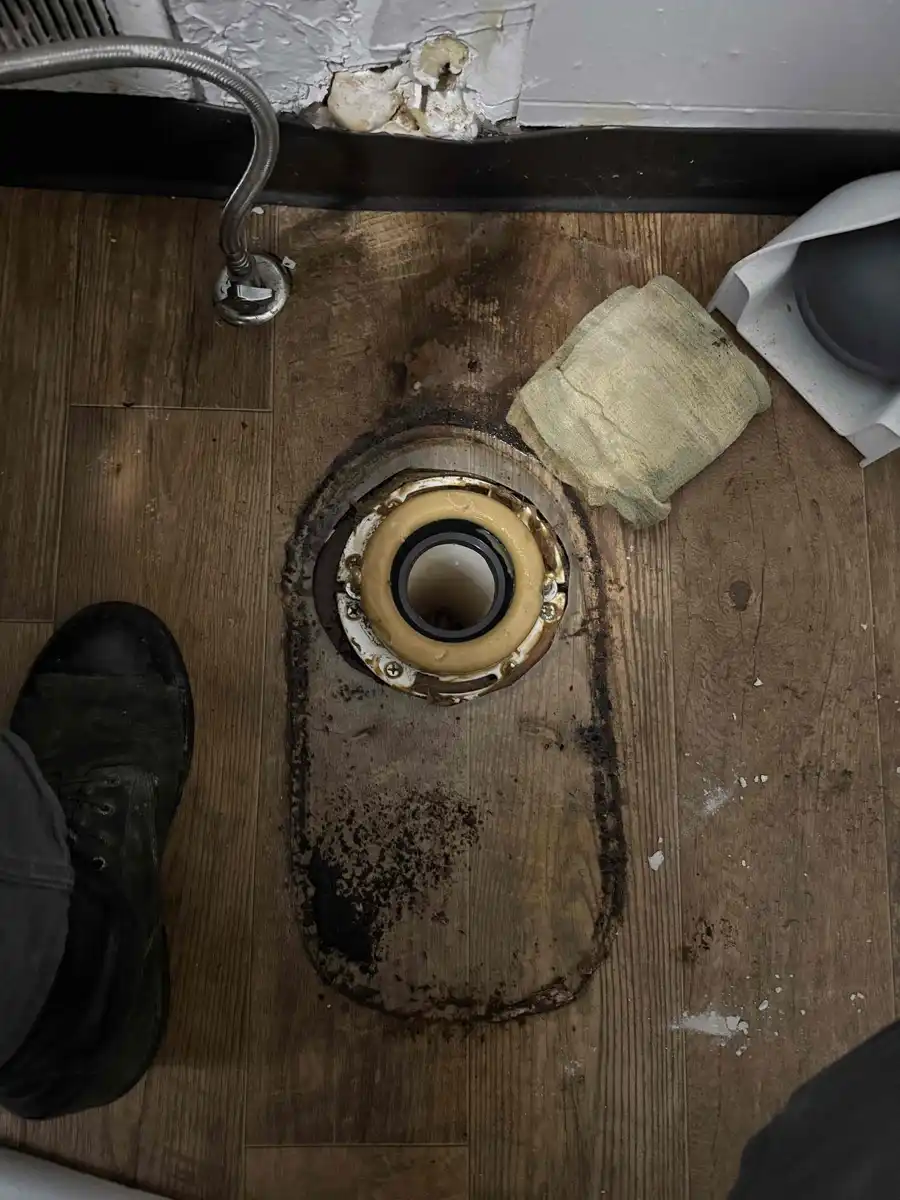 Toilet removed from the floor showing the closet flange and a new wax ring seated in it, with a dark stain in the shape of the toilet base on the vinyl plank floor