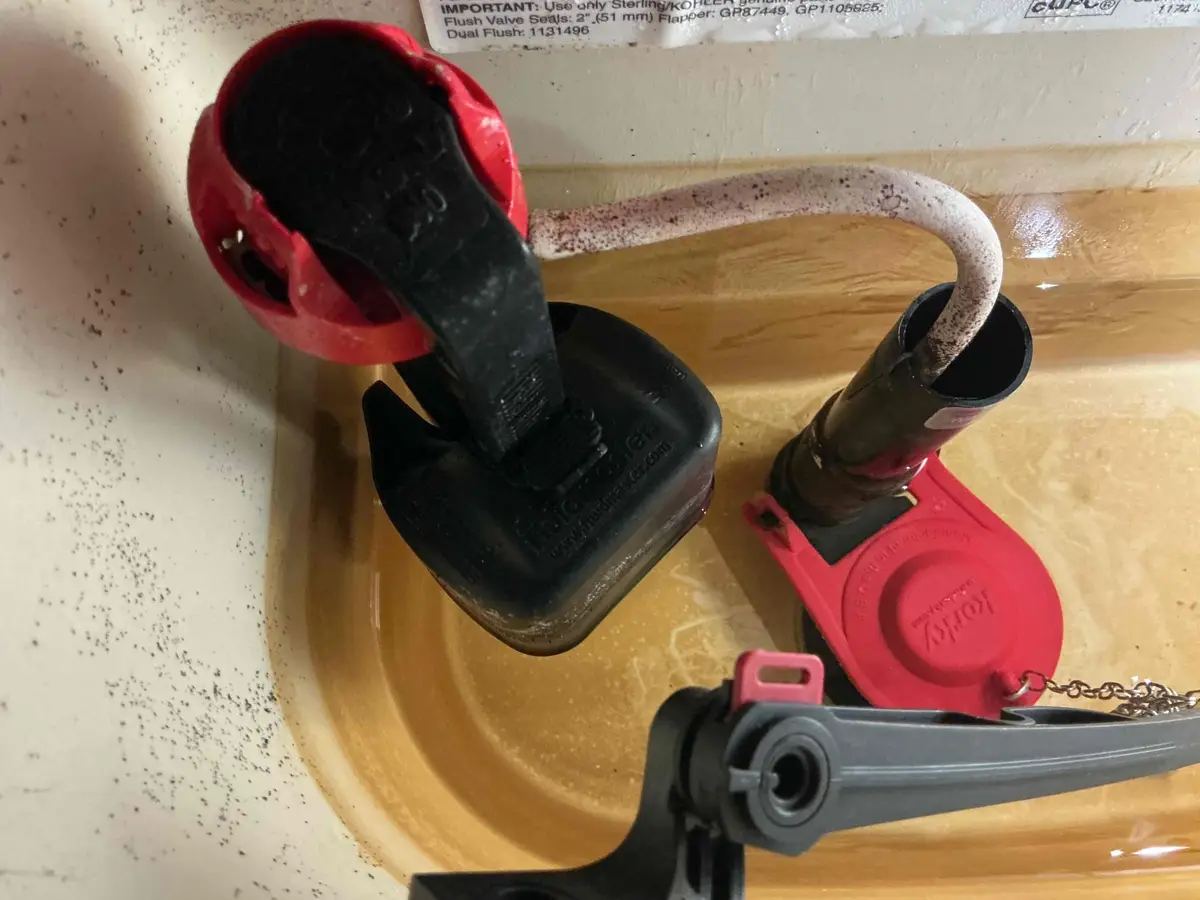 Close-up inside a toilet tank showing a red Korky flapper and fill valve with heavy mineral deposits and discolored water on the tank walls