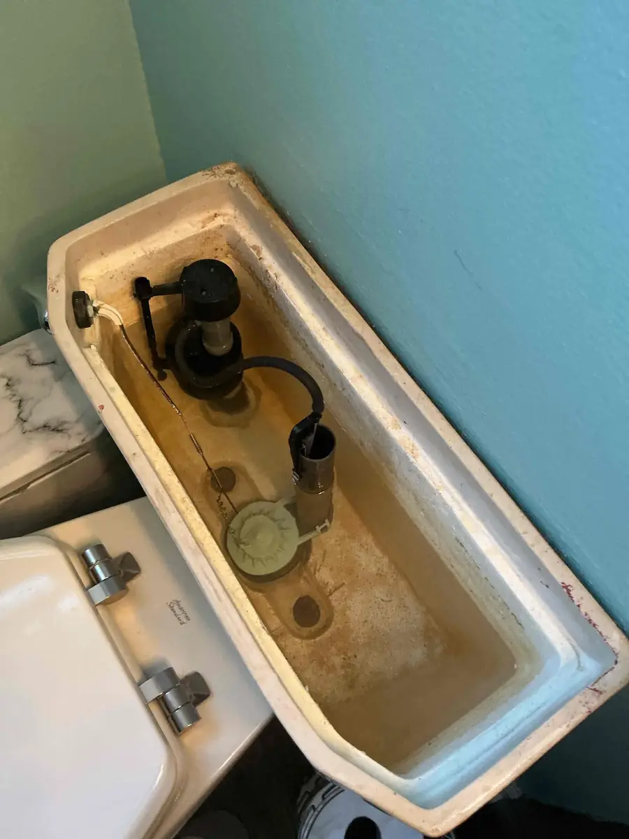 Inside of a residential toilet tank showing the fill valve on the left, flapper at the bottom center, and overflow tube on the right, with visible mineral staining on the tank walls