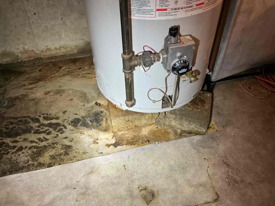 Failing gas water heater we removed, weeping from the tank body with rust staining spreading across the bare basement floor, no drain pan underneath