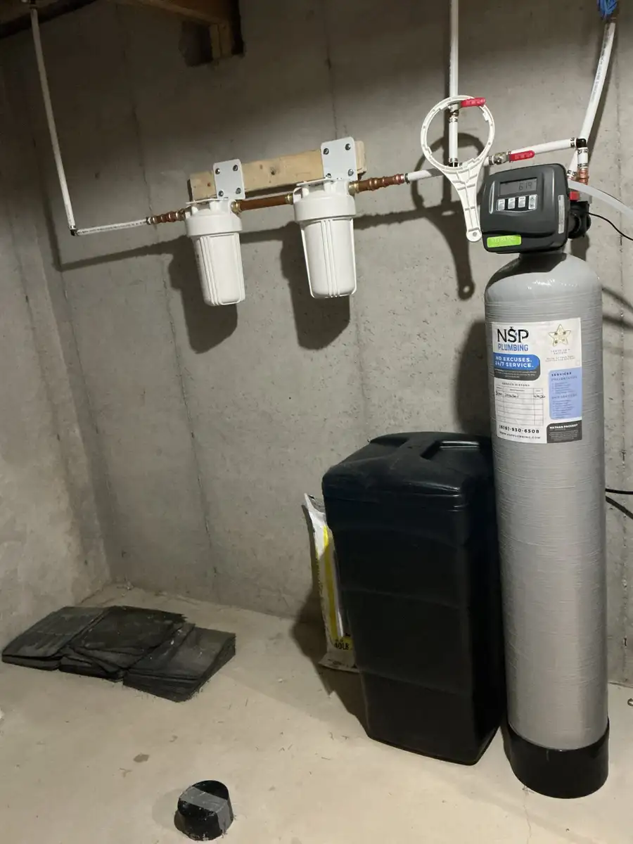 Water softener with NSP Plumbing service sticker, sediment filter canisters, and brine tank installed in a Grand Rapids basement