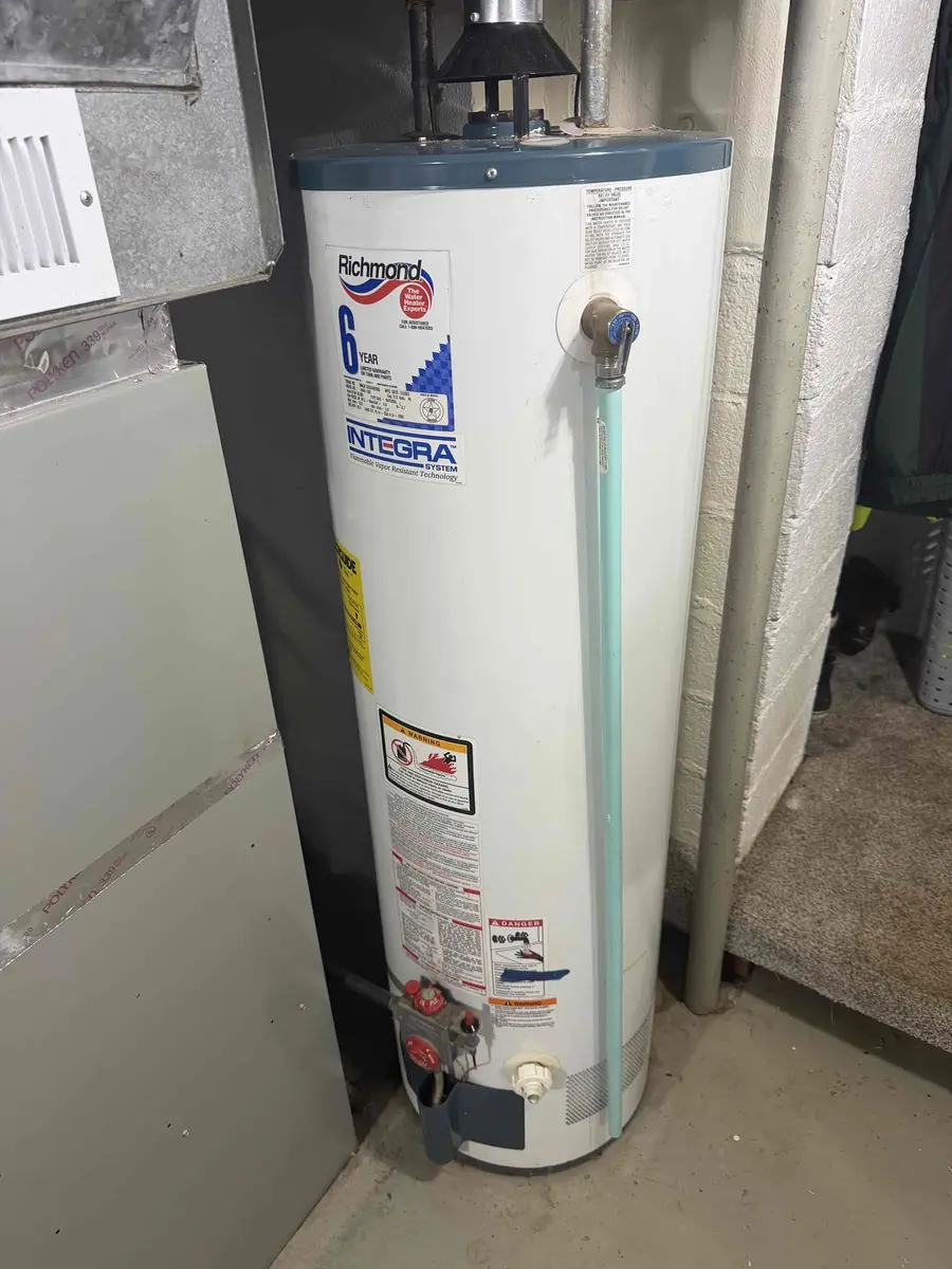 A gas water heater in a basement utility closet with the gas control valve, flue pipe, and temperature and pressure relief valve visible