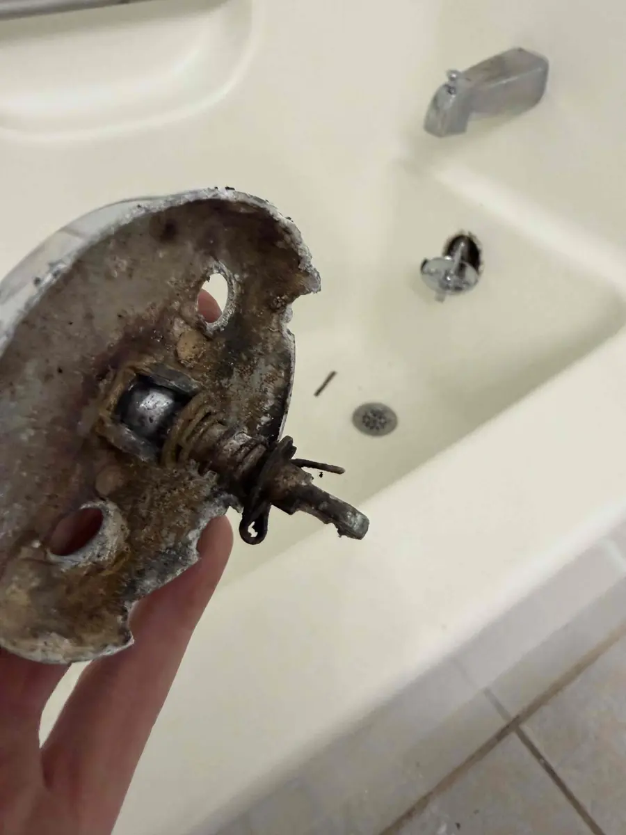 Hand holding a tub overflow plate with a crusted trip lever mechanism attached